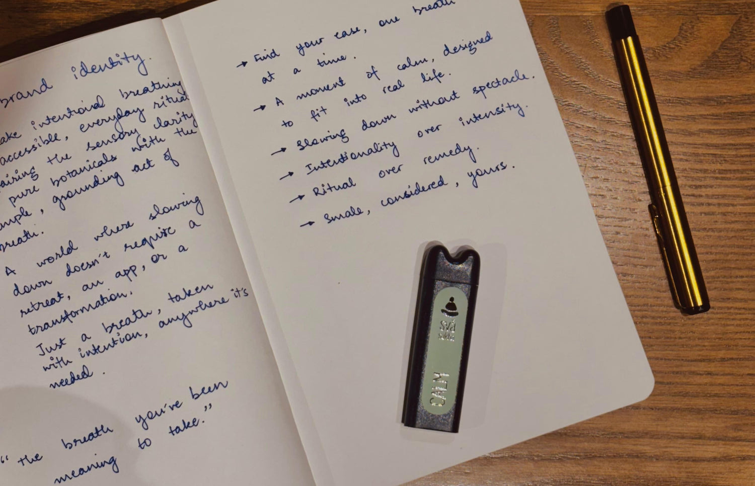 Svāroma aromatherapy inhaler placed on an open notebook with handwritten brand reflections on a wooden desk, next to a gold pen – calm, intentional workspace aesthetic